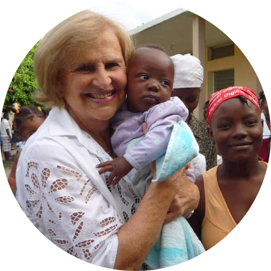 Dra. Zilda Arns segurando um bebê, sorrindo ao lado de mulheres em uma comunidade